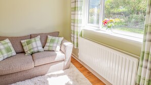 A cozy living room with a beige sofa, plaid cushions, a white fluffy rug, and a window with a view of a garden and hills. - Cam O'r Fechan Annexe (Llangollen)