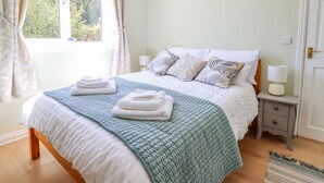 A neatly made bed with a blue quilt, white pillows, and folded towels on a wooden bed frame. There is a bedside table with a lamp and a small drawer. - Cam O'r Fechan Annexe (Llangollen)