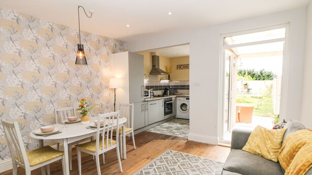 A modern kitchen with white cabinets, a dining area with a white table and chairs, and a grey sofa with yellow cushions.