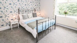 A bedroom with a metal bed frame, a white nightstand, a bedside lamp, and a window with a view of greenery.