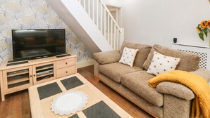 A living room with a TV on a wooden stand, a sofa with patterned cushions, a staircase, and a wall with floral wallpaper.