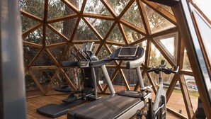 A gym with treadmills and exercise bikes, wooden flooring, and a large window with a view of trees. - Bento Box Vintage Family Villa & Hot Pool (Vilamoura)