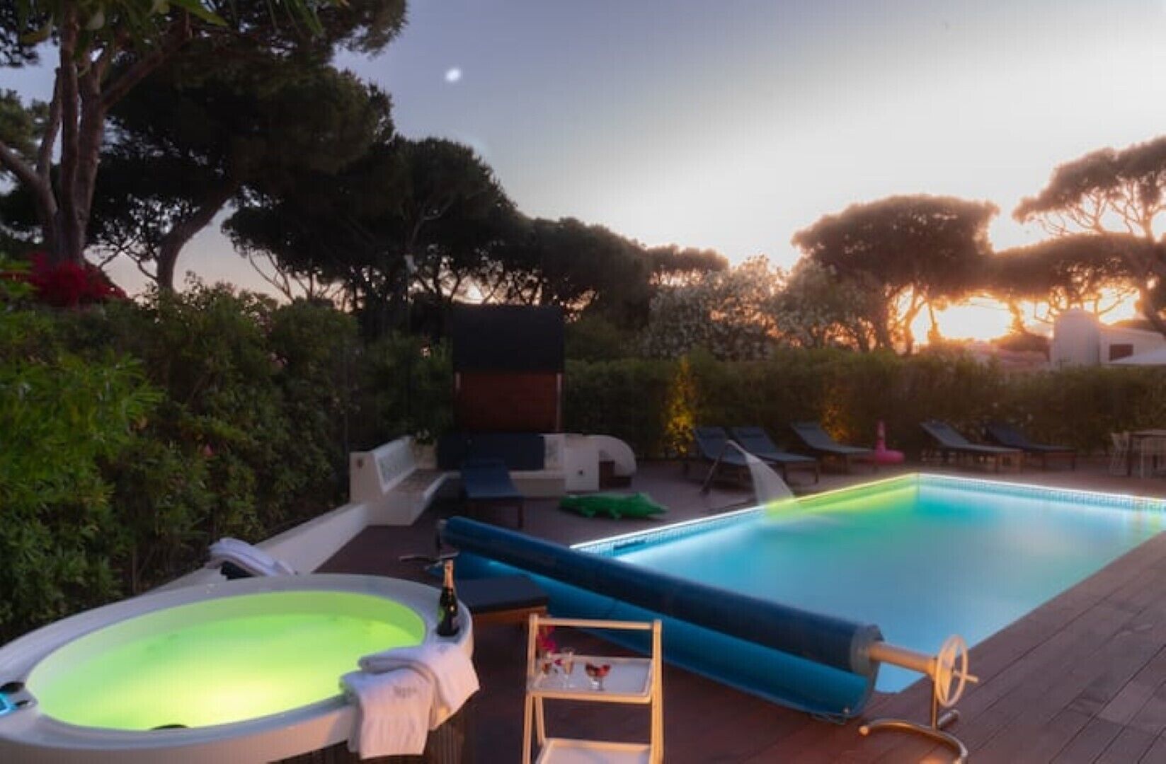 A swimming pool with lounge chairs and a hot tub, surrounded by trees and a sunset in the background.