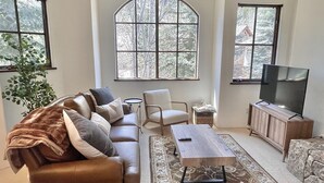 A living room with a brown leather sofa, a wooden coffee table, a TV on a wooden stand, and a large arched window. - JUST LISTED! Stylish townhome in West Ketchum -- private hot tub, close to bike path, river, ski lifts! (Ketchum)