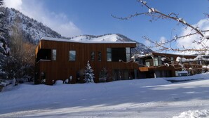 A modern wooden building with large windows, surrounded by snow-covered ground and trees, with a mountain in the background. - JUST LISTED! Stylish townhome in West Ketchum -- private hot tub, close to bike path, river, ski lifts! (Ketchum)