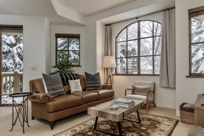 Relax in this inviting living room with a leather sofa, coffee table, and side table, surrounded by large windows and a potted plant.
