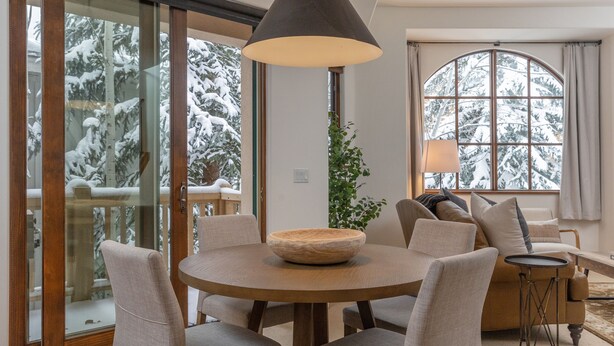 A cozy living room with a round wooden table, beige sofas, and a large window with a view of snow-covered trees. - JUST LISTED! Stylish townhome in West Ketchum -- private hot tub, close to bike path, river, ski lifts! (Ketchum)