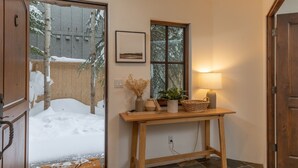 A wooden door leading to a snow-covered outdoor area with a window and a table with a lamp, vase, and basket. - JUST LISTED! Stylish townhome in West Ketchum -- private hot tub, close to bike path, river, ski lifts! (Ketchum)