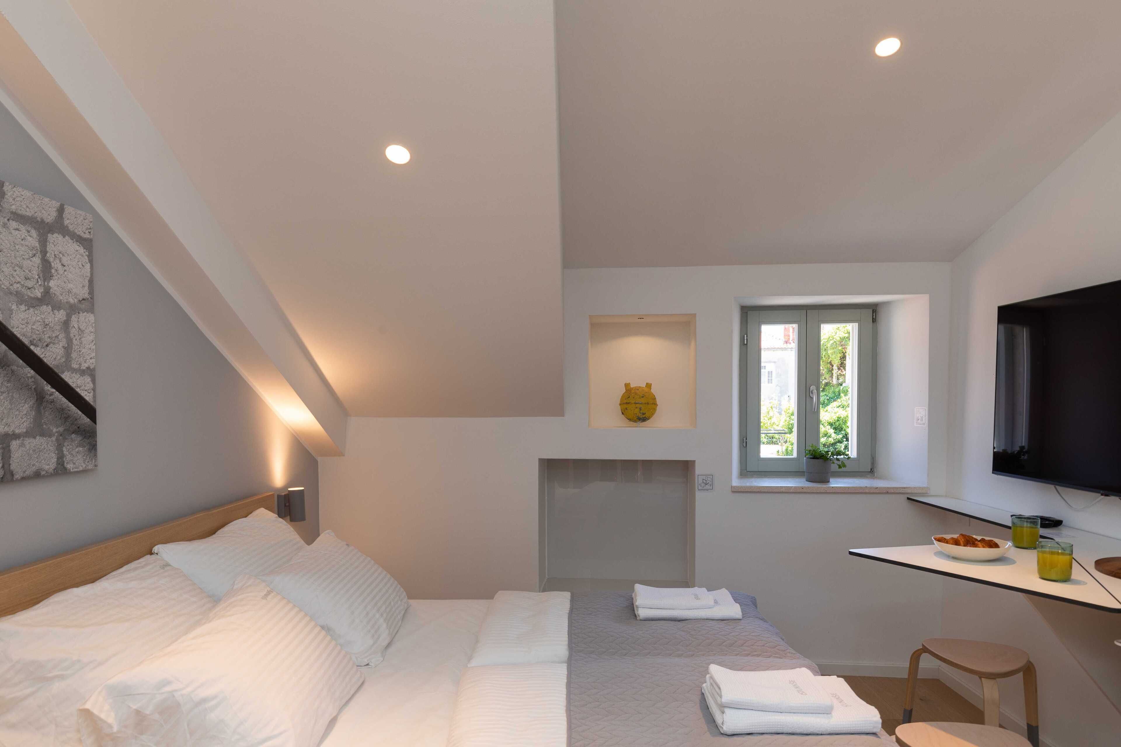 A bedroom with a bed, a desk with a chair, a television, and a window with a plant.