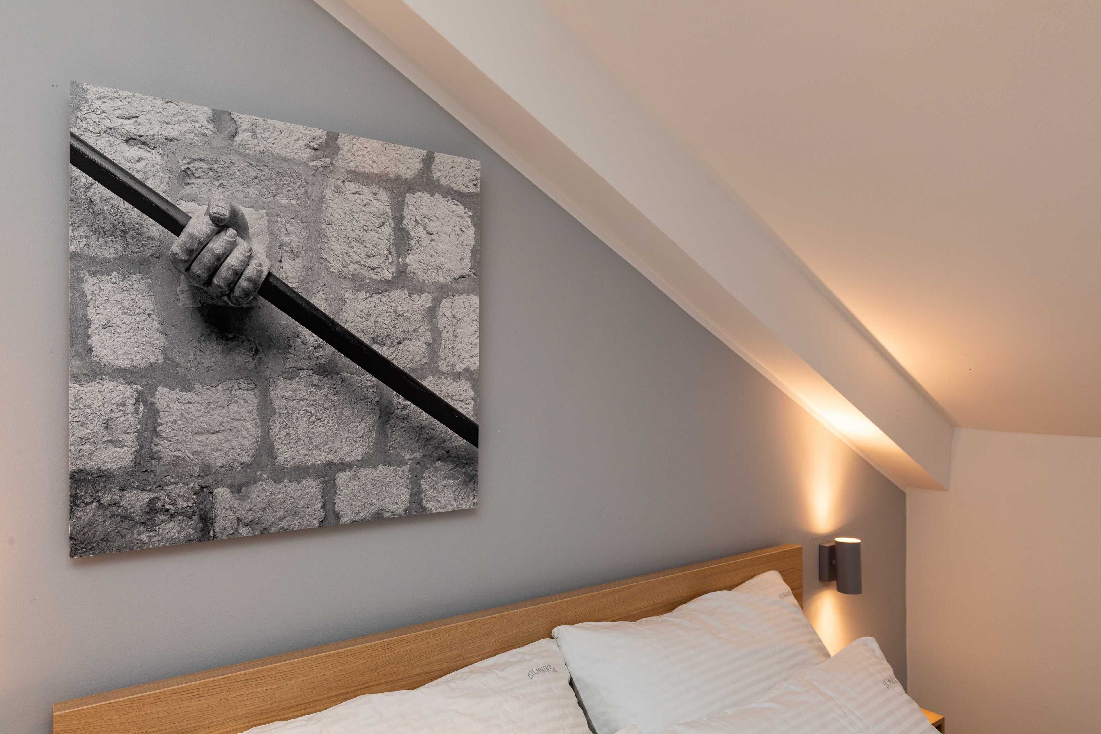 A bedroom with a bed, a wall-mounted artwork of a hand holding a pole, and a wall-mounted light fixture.