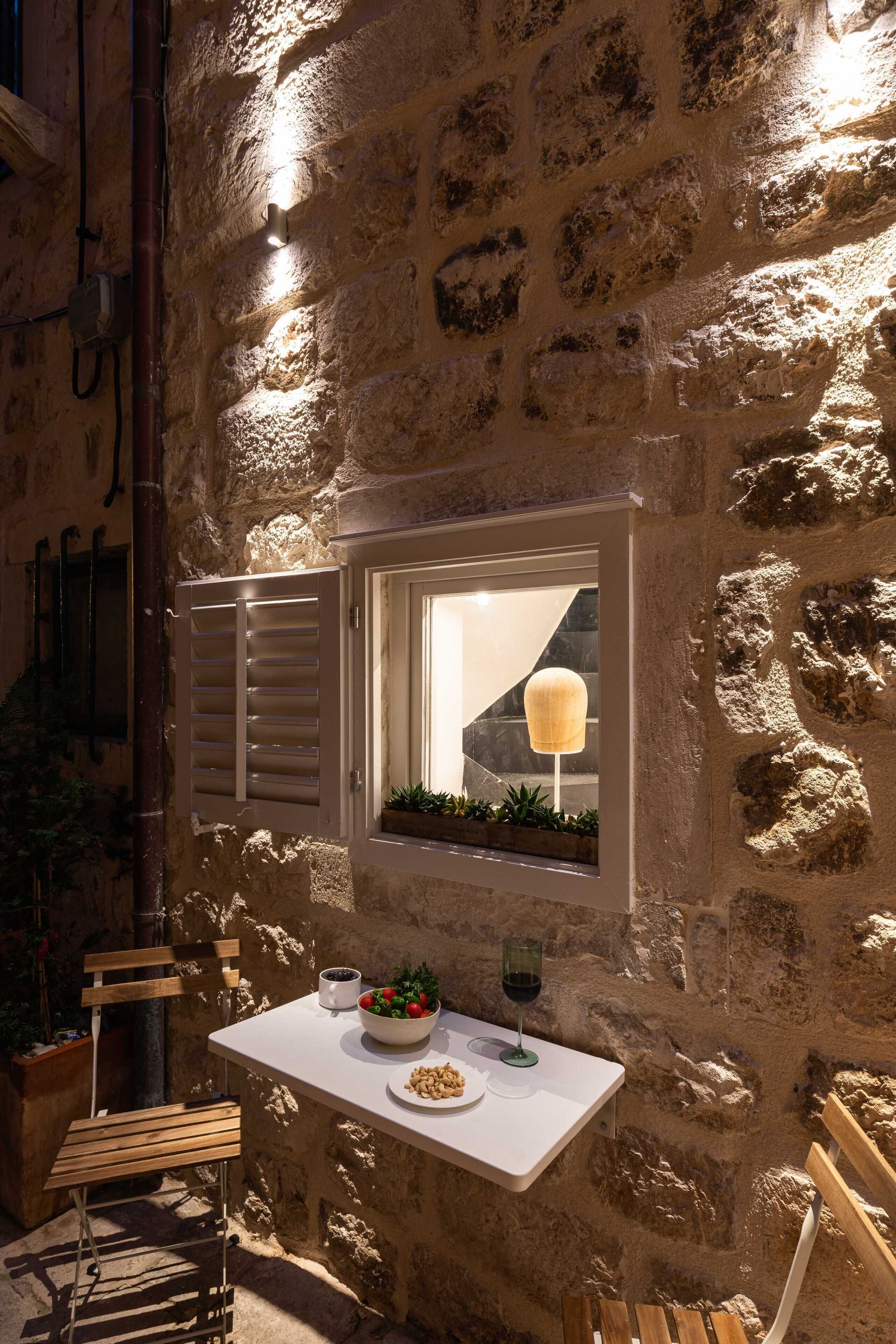 A stone wall with a window, a table with a bowl of fruit and a glass of wine, and wooden chairs.
