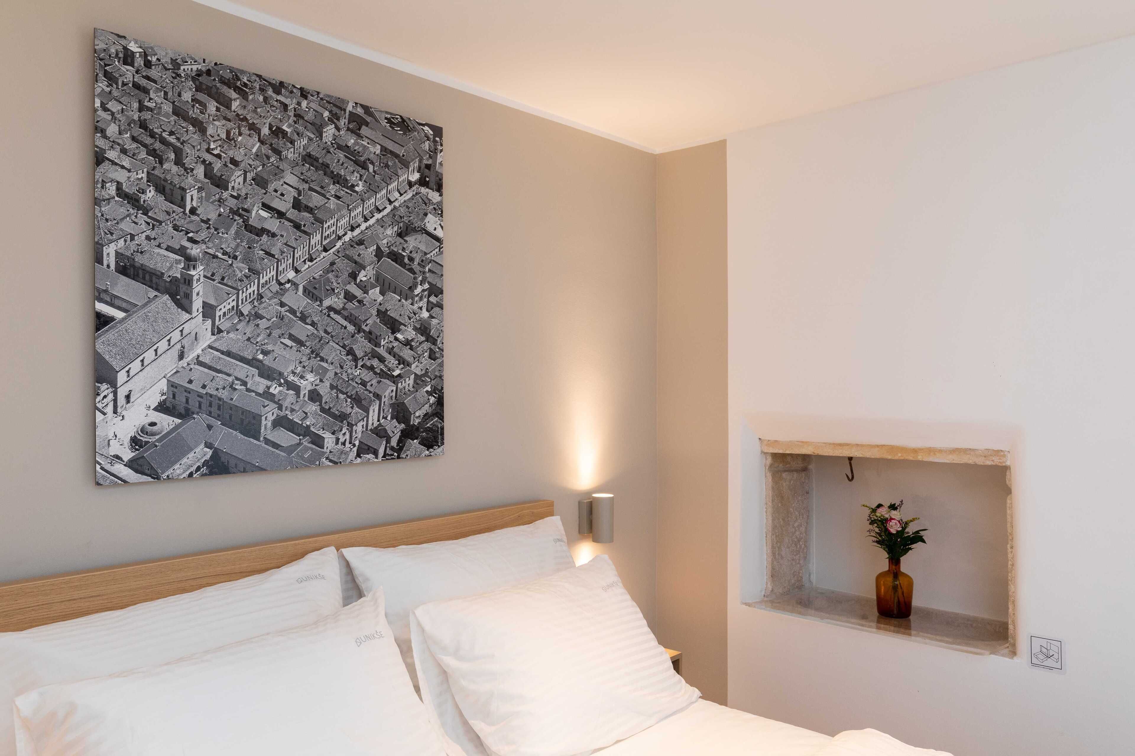 A hotel room with a large black and white cityscape painting, a wooden headboard, white bedding, a bedside lamp, and a small window with a vase.