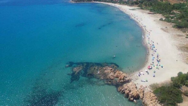 Vista aérea de uma praia costeira com águas azuis cristalinas, areia dourada e alguns edifícios dispersos ao fundo.