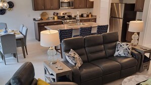 A modern living room with a black leather sofa, a dining area with a table and chairs, and a kitchen with wooden cabinets and stainless steel appliances. - 3/2 IN THE VILLAGE OF ST. CATHERINE GAS GOLF CART, CLOSE TO SAWGRASS GROVE (Wildwood)