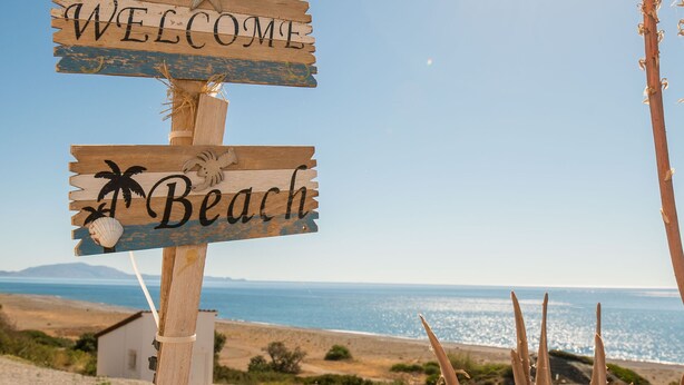 Un panneau indicateur en bois portant les inscriptions « BIENVENUE » et « PLAGE », placé sur un fond de paysage côtier avec un ciel dégagé et un soleil brillant.