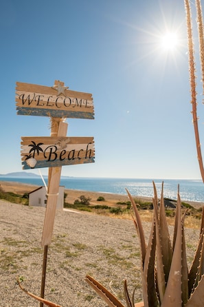 A wooden signpost with 'WELCOME' and 'Beach' written on it, set against a coastal backdrop with the ocean and a clear sky. - Παραλιακη, εξοχικη κατοικια μπροστα στην θαλασσα. (Ρόδος)