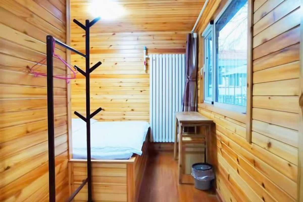 Une petite pièce de cabane en bois comprenant un lit, un porte-manteau, une petite table et une fenêtre.