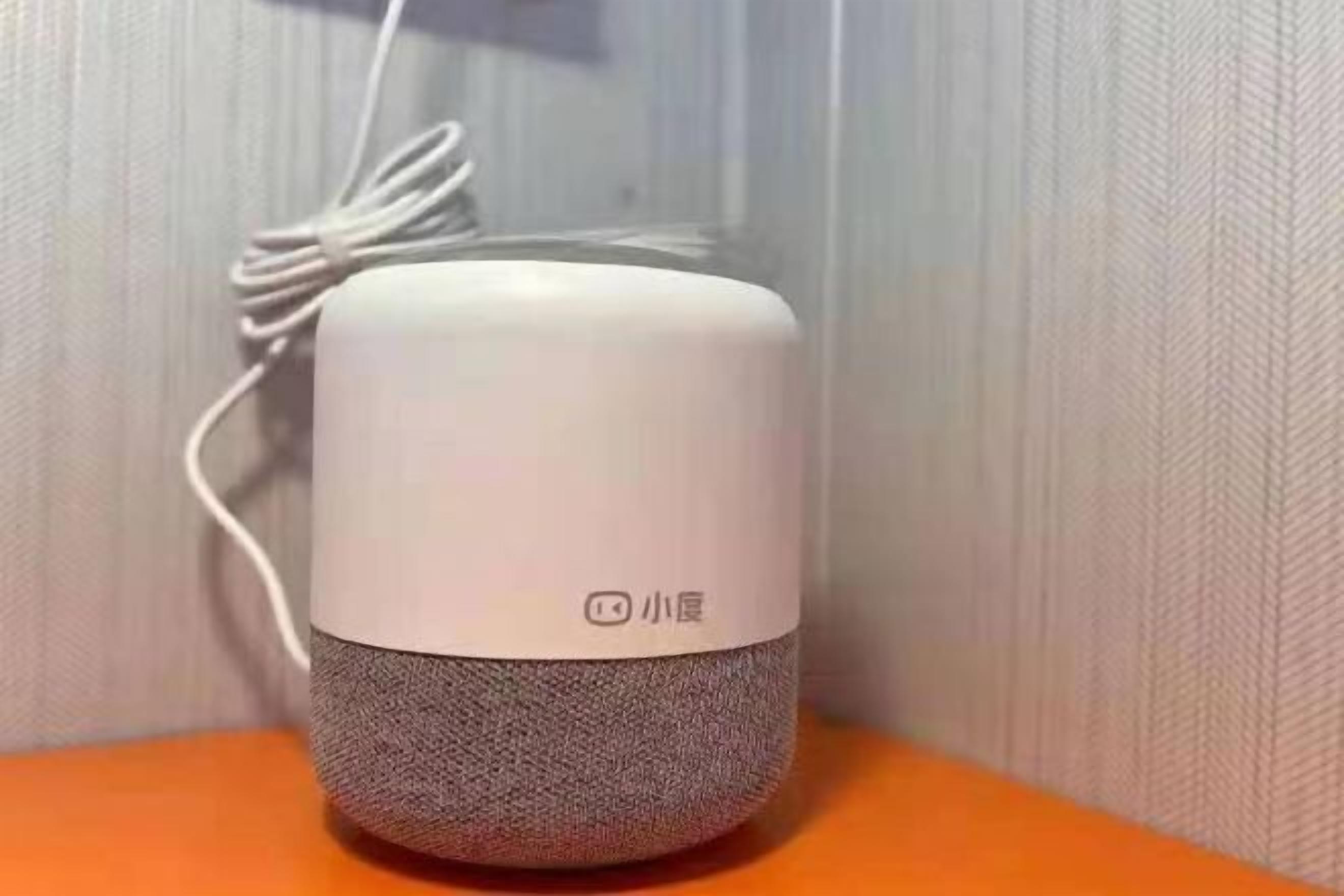 Un petit appareil cylindrique blanc, doté d’une base texturée, qui ressemble probablement à un haut-parleur ou à un appareil audio, est posé sur une surface orange, devant un mur texturé.