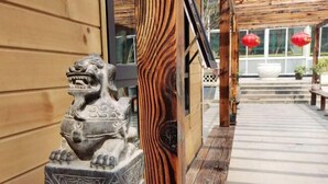 Une statue de lion en pierre, posée sur un piédestal, un mur en bois à lattes et une terrasse en bois décorée de lanternes rouges.