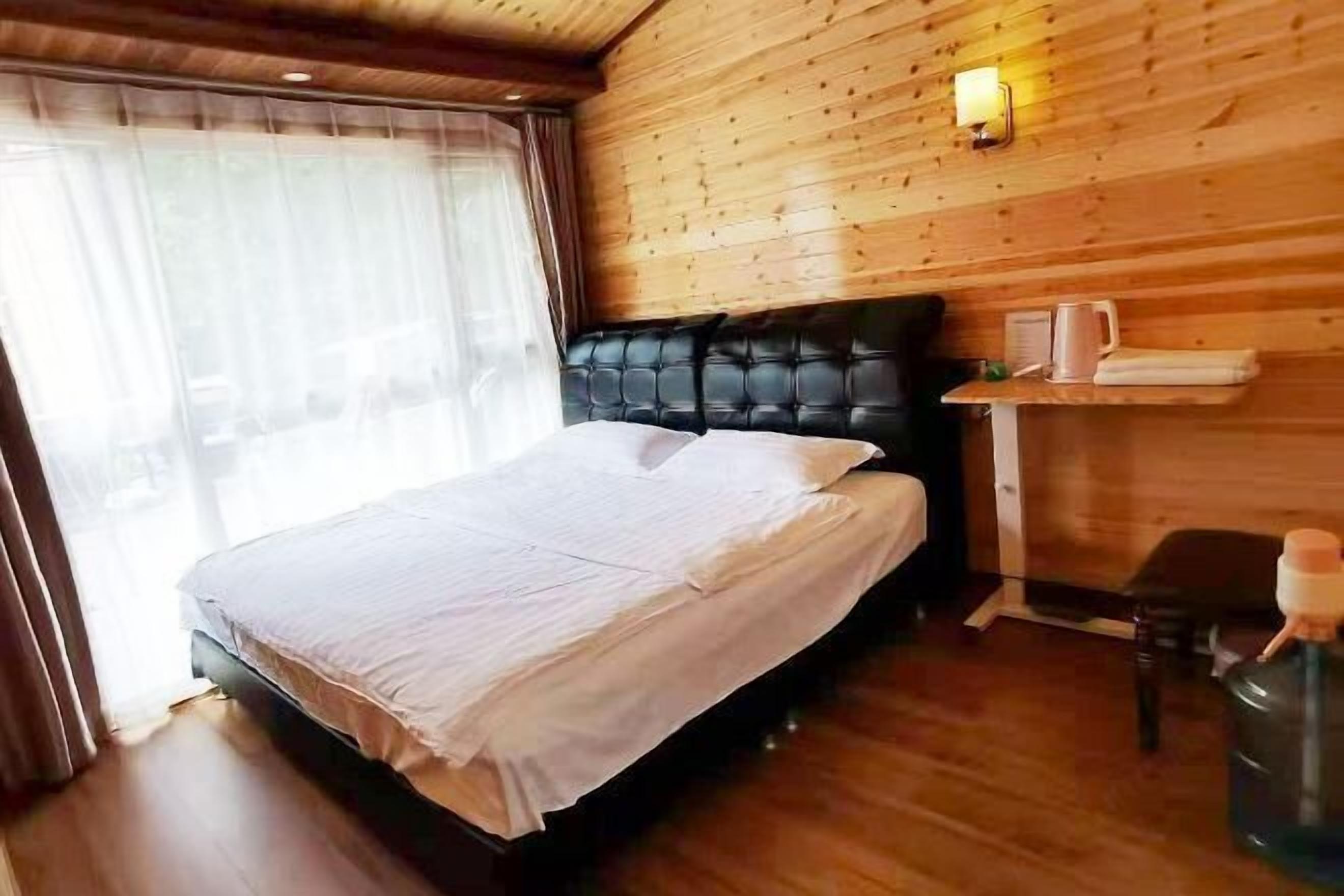 Un dormitorio con cama, paredes de madera, ventana con cortinas, mesita de noche con hervidor y una silla.