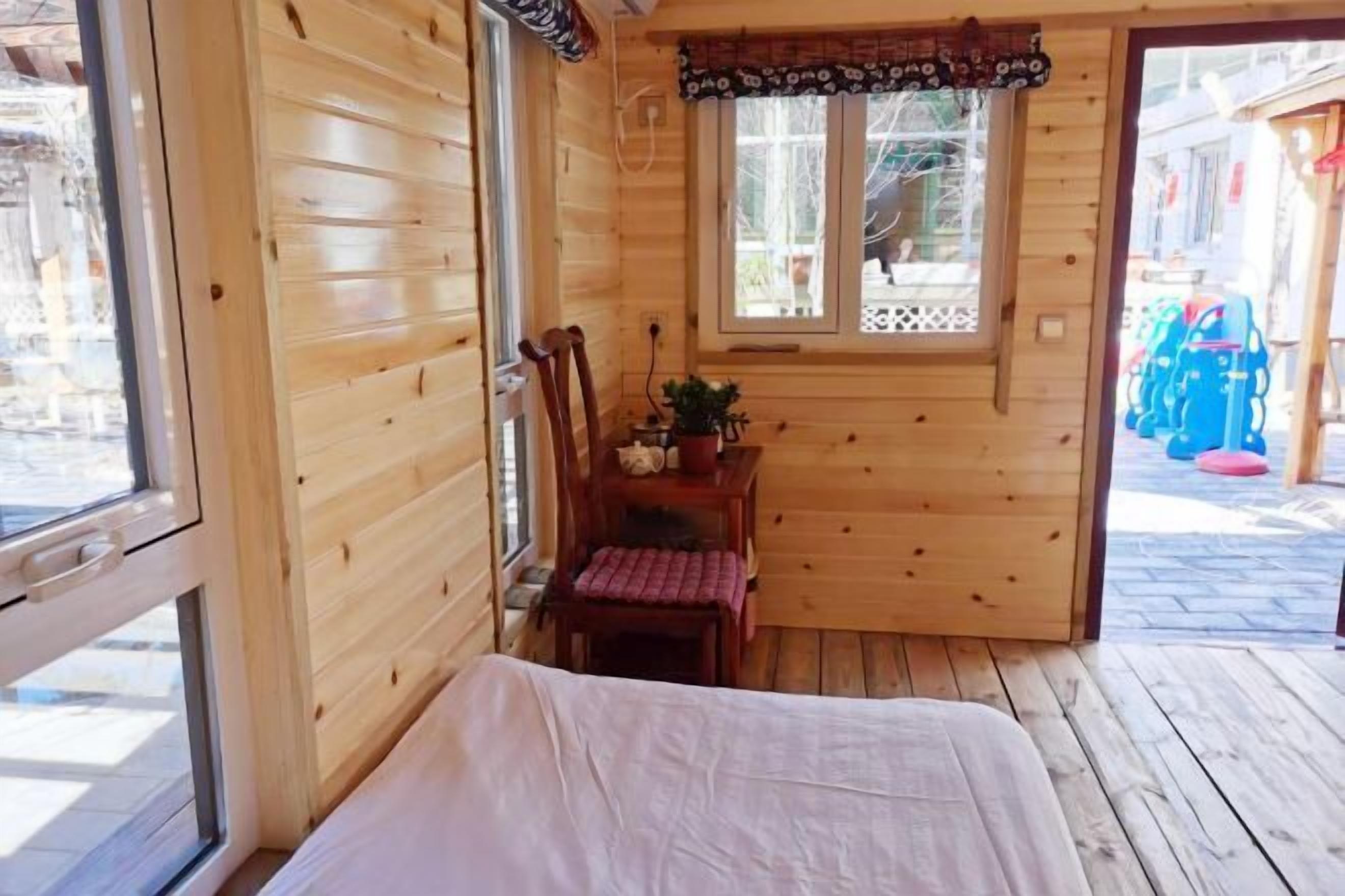 A wooden cabin room with a bed, a window, a small table with a plant, and a chair.
