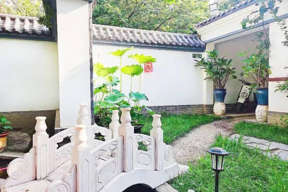 Une porte de style chinois traditionnel, ornée de sculptures complexes, un sentier menant à un bâtiment doté d’un toit en tuiles et des plantes en pot.