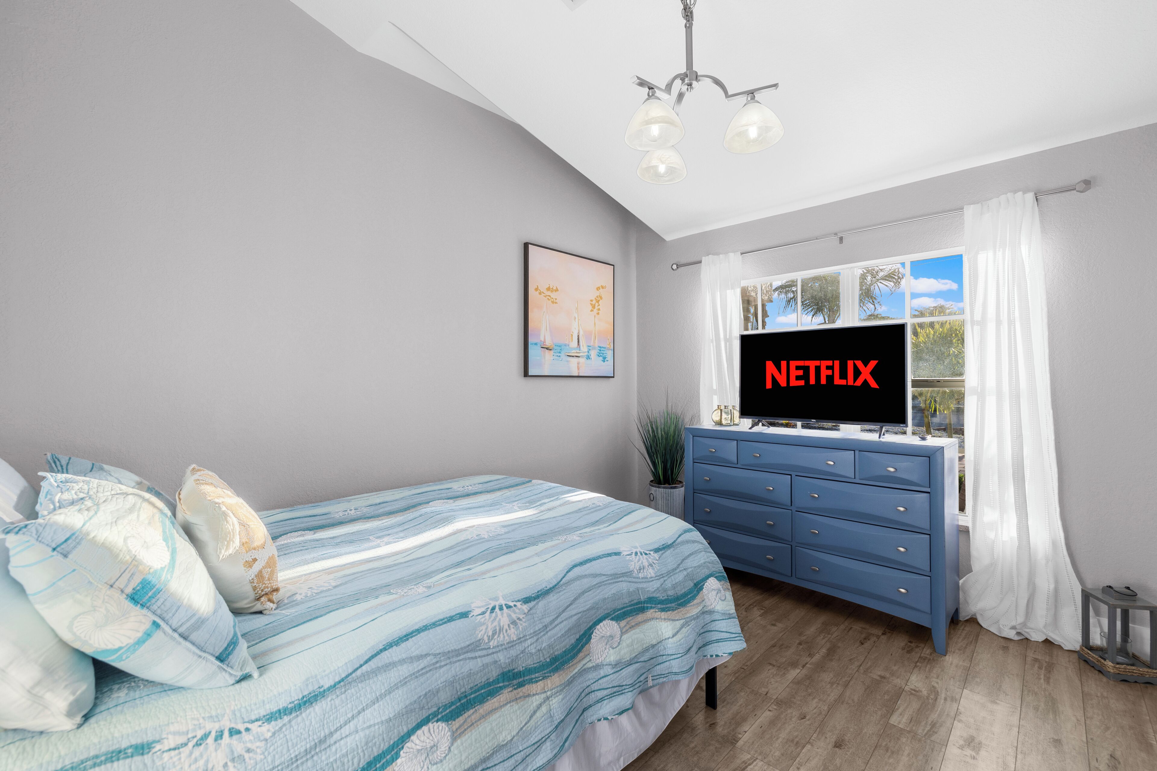 Une chambre à coucher avec un lit, une commode, un téléviseur affichant le logo « NETFLIX », une fenêtre avec des rideaux et un luminaire au plafond.