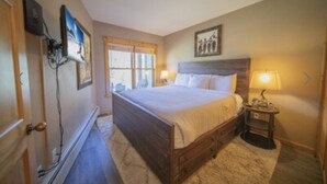 A bedroom with a wooden bed, a nightstand with a lamp, a window with curtains, and a wall-mounted TV. - Keystone Ski area, Epic mountains views, Ground floor condo, short walk to lift  (Keystone)