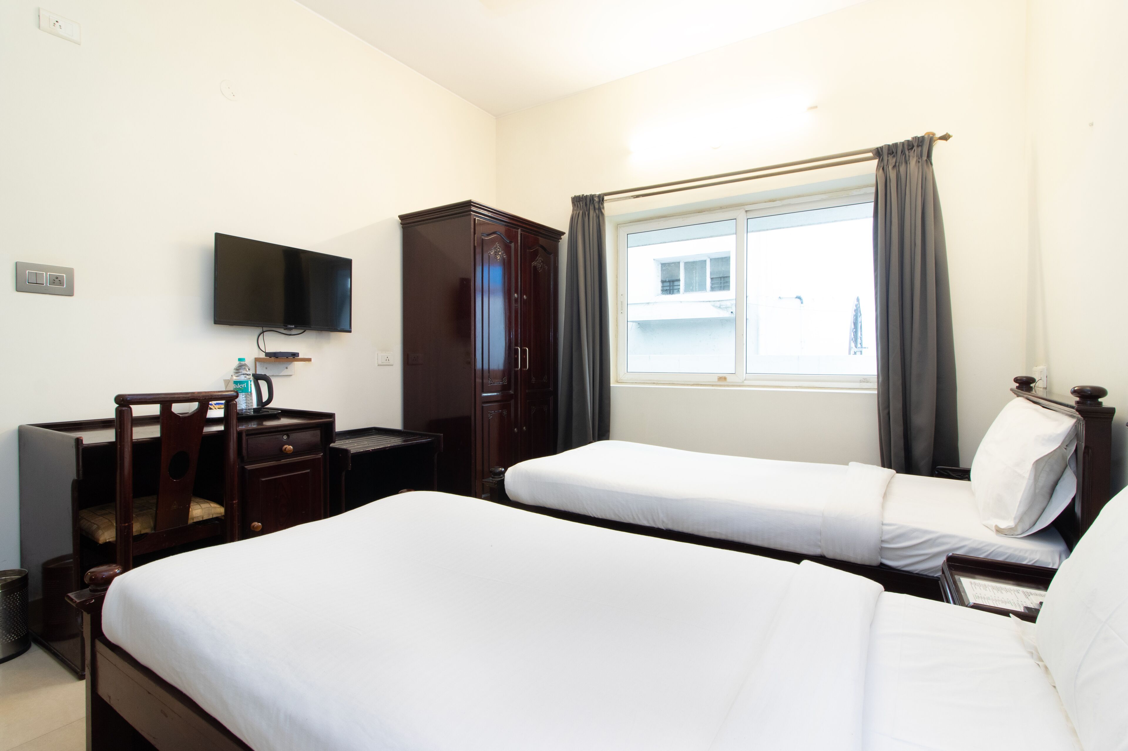A hotel room with two single beds, a desk with a chair, a television mounted on the wall, and a window with curtains.