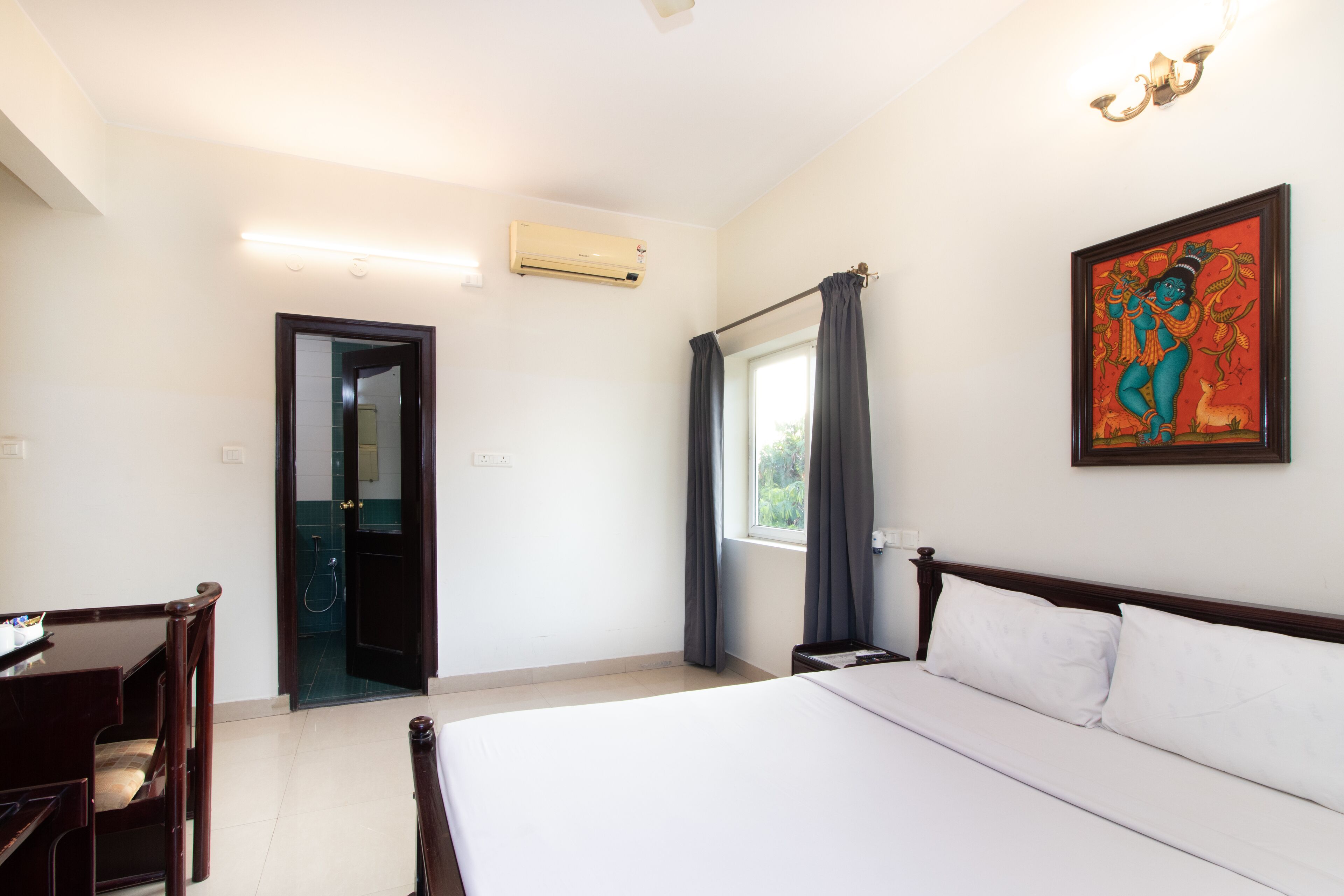 Une chambre d’hôtel comprenant un lit, un bureau, une chaise, une fenêtre avec des rideaux, un climatiseur et une œuvre d’art accrochée au mur.