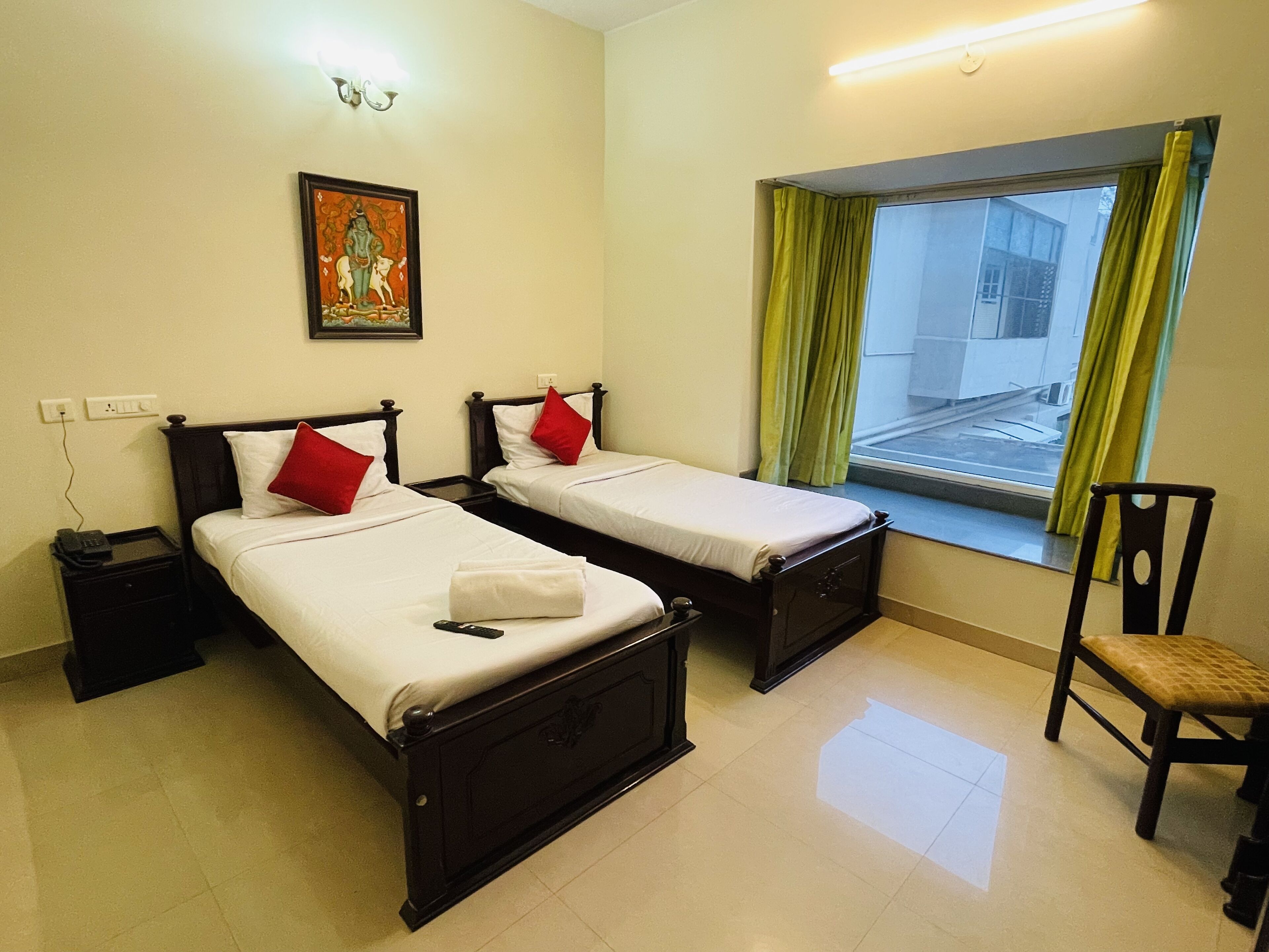 A room with two beds, a chair, a window with curtains, a wall-mounted painting, and a bedside table with a phone.