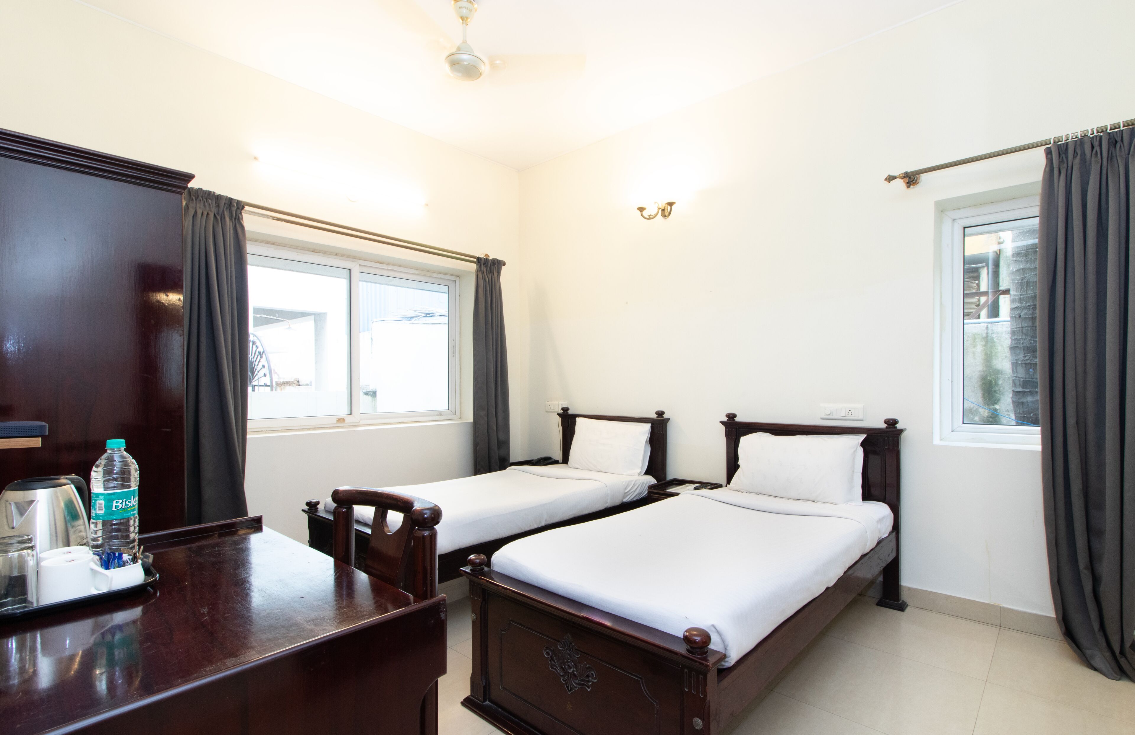 A hotel room with two single beds, a wooden desk with a water bottle, a coffee maker, and a small table.