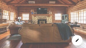 A log cabin interior with a stone fireplace, a large leather sofa, and a television mounted above it. -  Cabin By the Bay A unique authentic log cabin by the beach with fenced yard (Panacea)