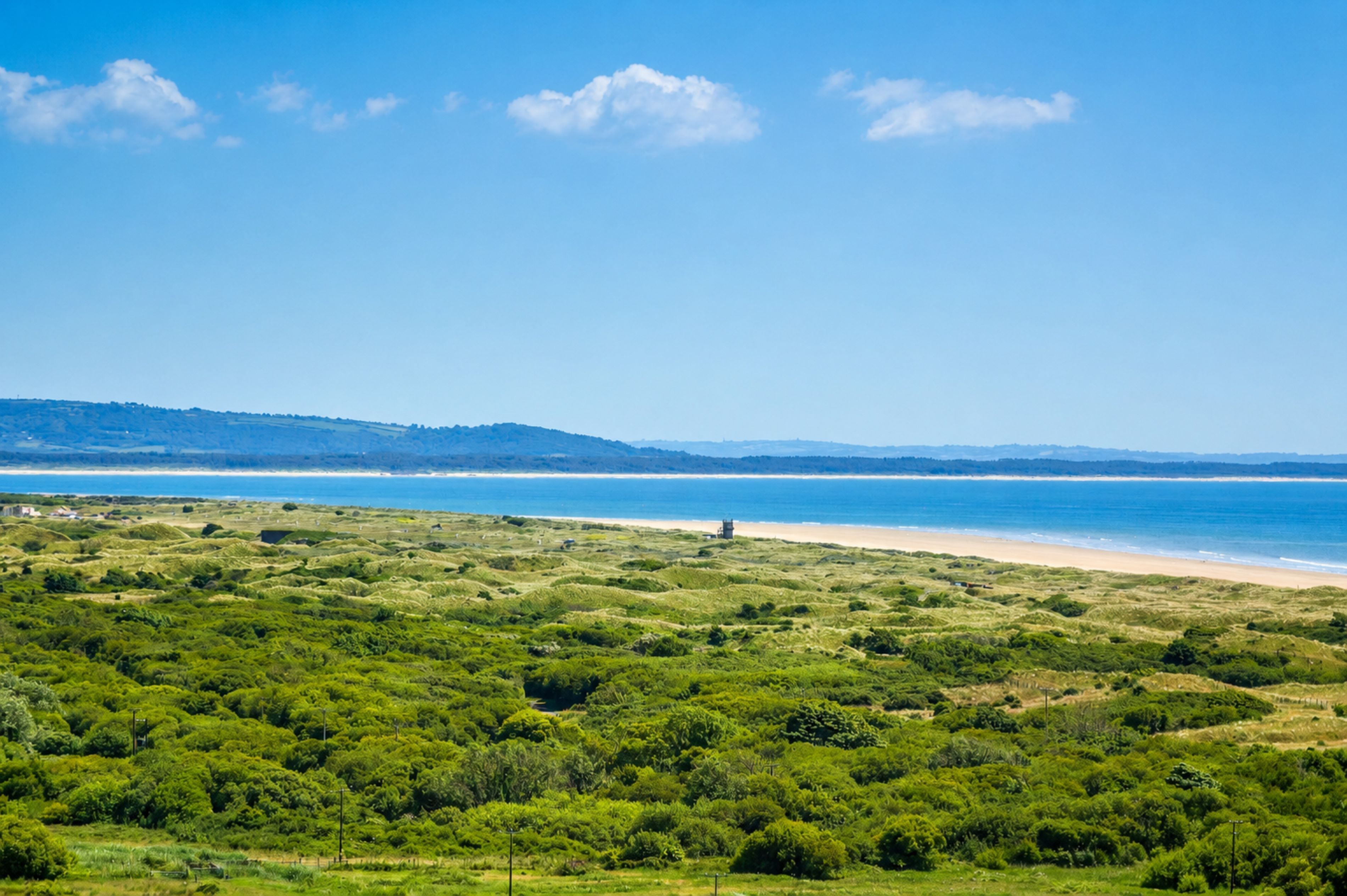 Een kustlandschap met een strand, glooiende heuvels en een heldere lucht.