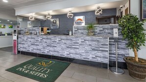 A reception desk with a stone facade, a sign reading "Quality in & Suites," and a potted plant to the right. - Suburban Studios Rock Hill Northeast I-77 (Rock Hill)