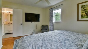 Ein Schlafzimmer mit einem Bett, einem Deckenventilator, einem an der Wand befestigten Fernseher, einem Fenster mit Vorhängen und einer Tür zu einem Badezimmer.