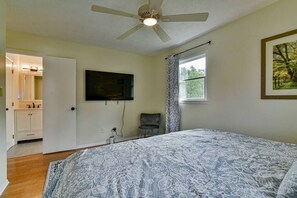 Une chambre à coucher comprenant un lit, un ventilateur de plafond, un téléviseur fixé au mur, une fenêtre avec des rideaux et une porte donnant sur une salle de bain.