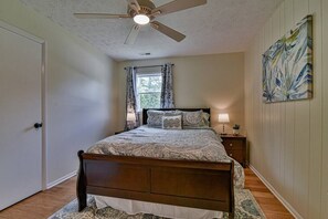 Quarto com cama, ventilador de teto, janela com cortinas, mesa de cabeceira com abajur e um quadro na parede.