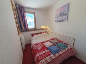 A small bedroom with a bed, a window with curtains, a wall-mounted lamp, and a painting.