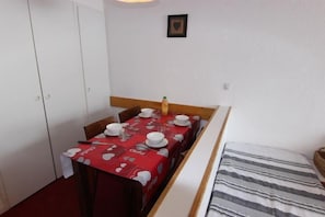 A small room with a dining table set for two, a bed with striped bedding, and a white wardrobe.