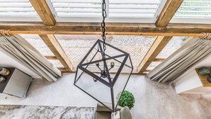 Ein Zimmer mit einer geometrisch gestalteten Pendelleuchte, Holzbalken und Blick auf einen Garten durch große Fenster.