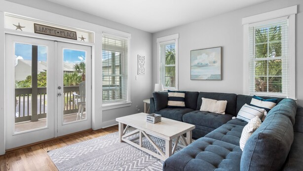 A living room with a grey sofa, a white coffee table, and a patterned rug. There are two windows with blinds, a door with a 'Happy Place' sign, and a painting on the wall.