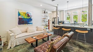 Ein modernes Wohnzimmer mit einer Couch, einem Couchtisch und Barhocker. Im Hintergrund ist eine Küche mit einer Bar und großen Fenstern zu sehen.