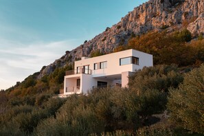 Ein modernes, zweistöckiges Haus mit Balkon, umgeben von dichter Vegetation und einer felsigen Hanglage.