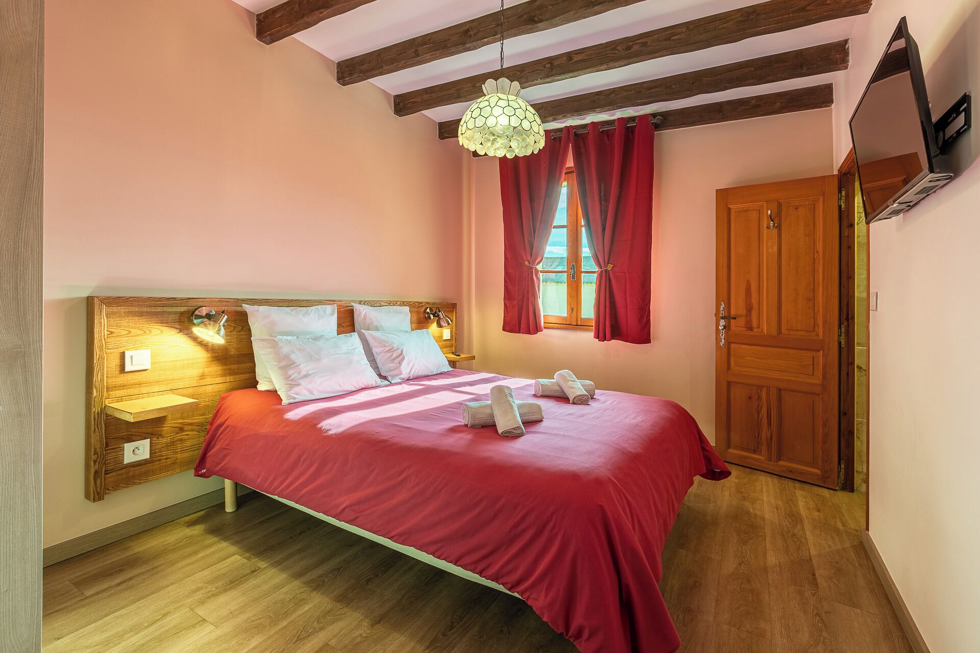 A bedroom with a wooden headboard, a bed with red bedding, a wooden floor, a window with red curtains, and a wooden door.