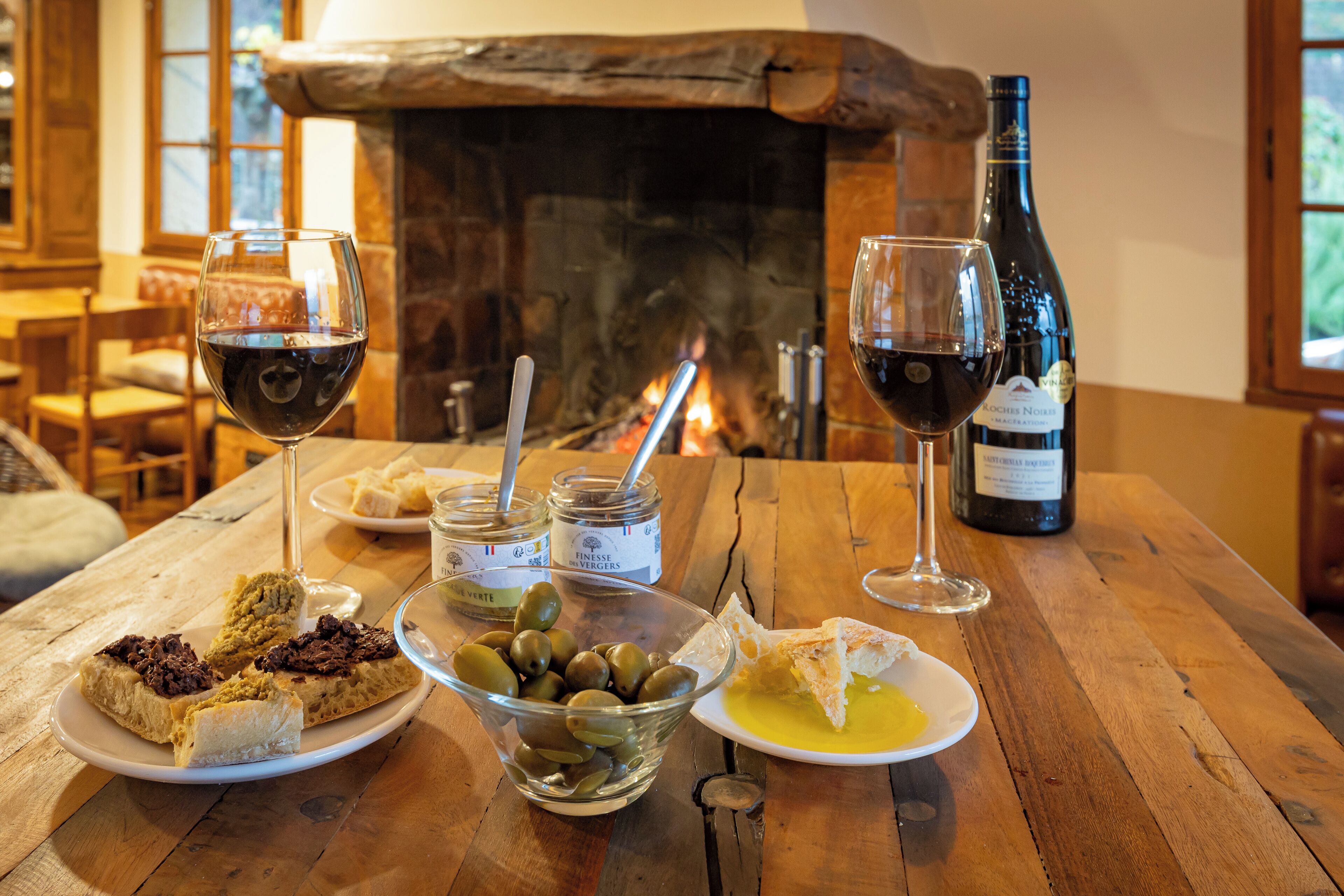 A wooden table with a bottle of wine, glasses, olives, bread, and a bowl of olives.