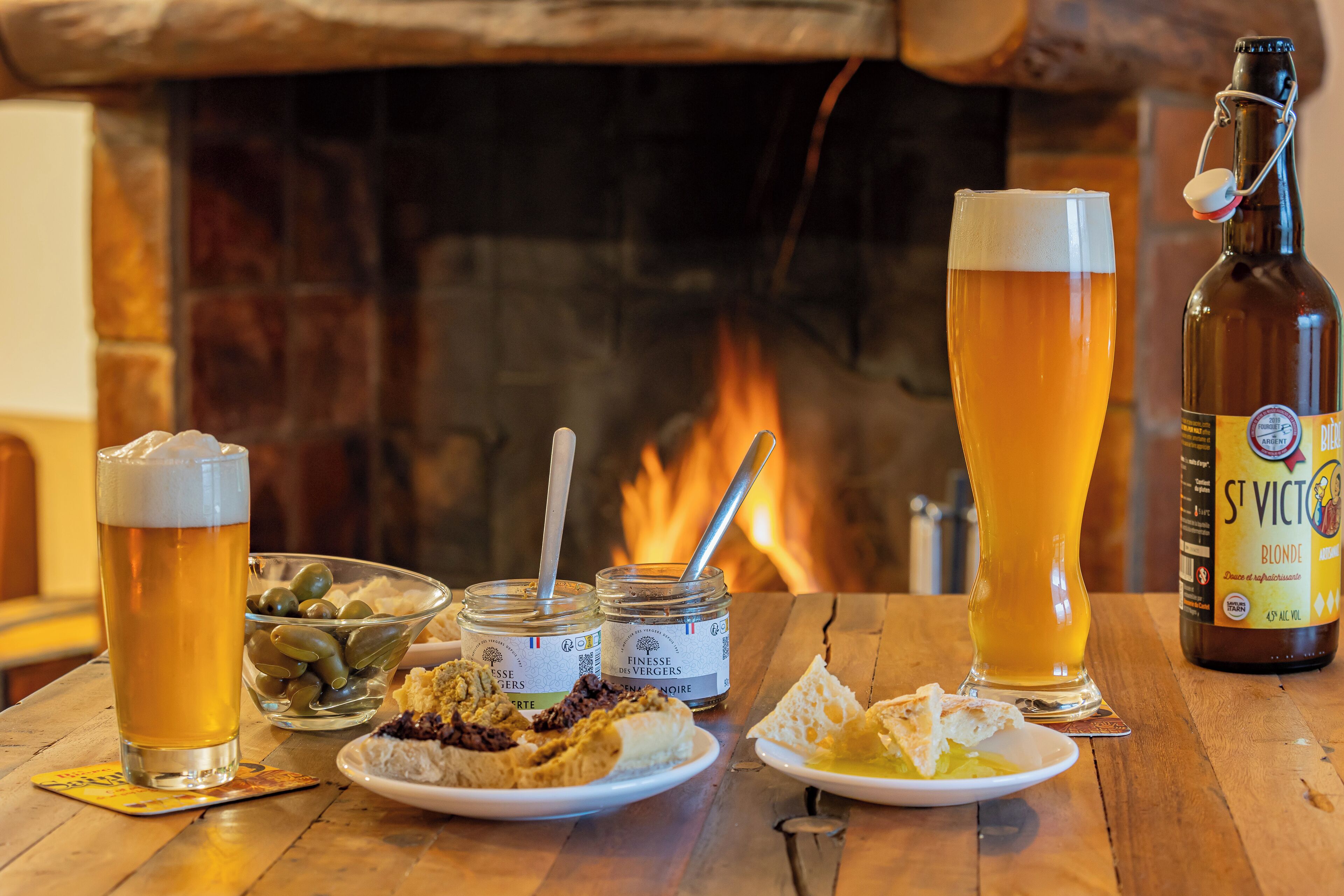 Una mesa con una botella de cerveza, dos vasos de cerveza, un cuenco de aceitunas y platos de comida.