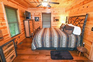 Un dormitorio de cabaña de madera con cama, cómoda, ventilador de techo y ventana con persianas.