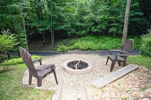 A circular seating area with a fire pit, surrounded by gravel, with two Adirondack chairs and a wooden bench.