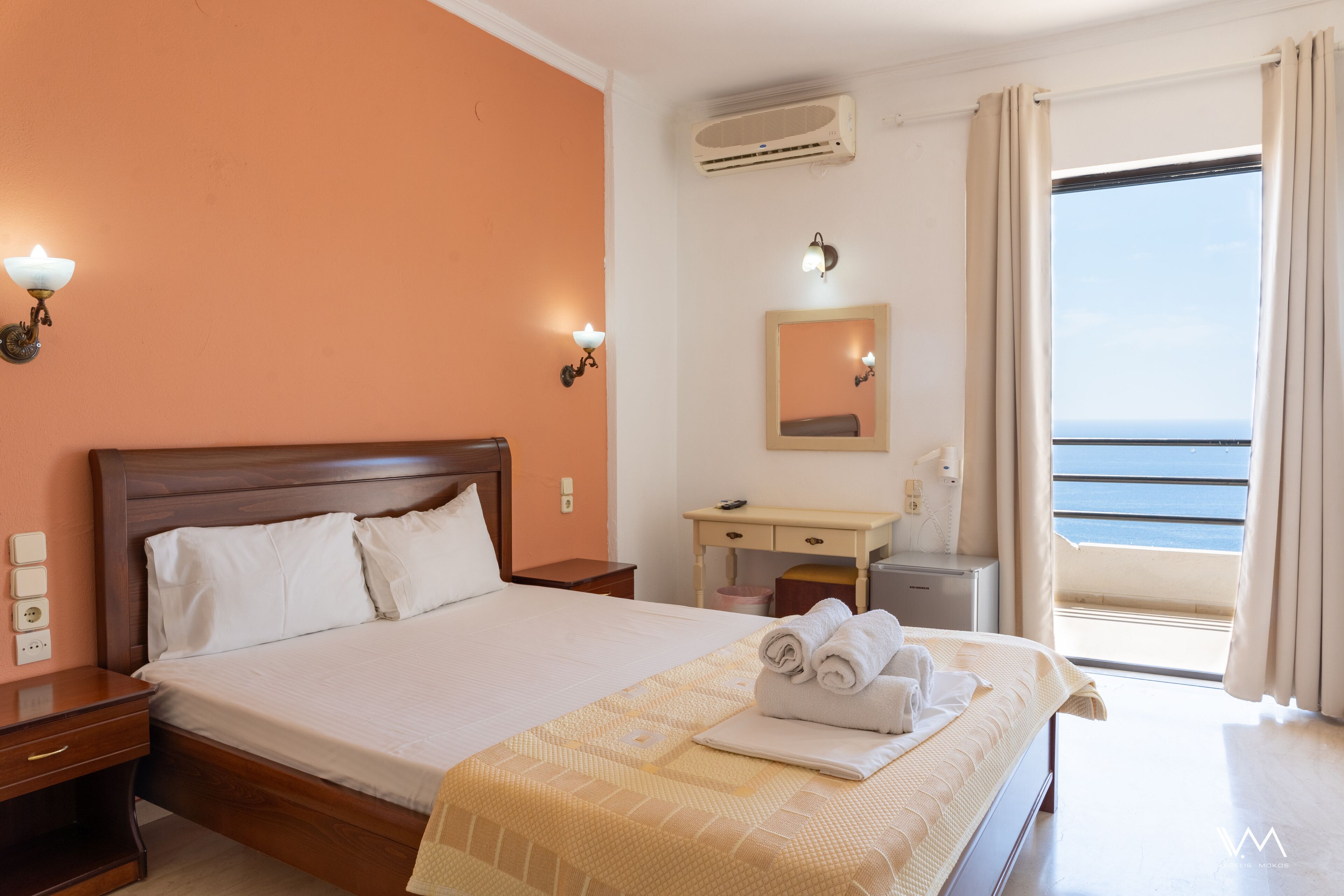 Quarto de hotel com cama, mesas de cabeceira, escrivaninha, ar condicionado, espelho e janela com cortinas.